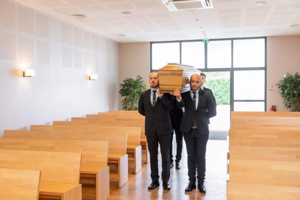 Porteurs en costume sombre portant un cercueil en bois lors d'une cérémonie funéraire.
